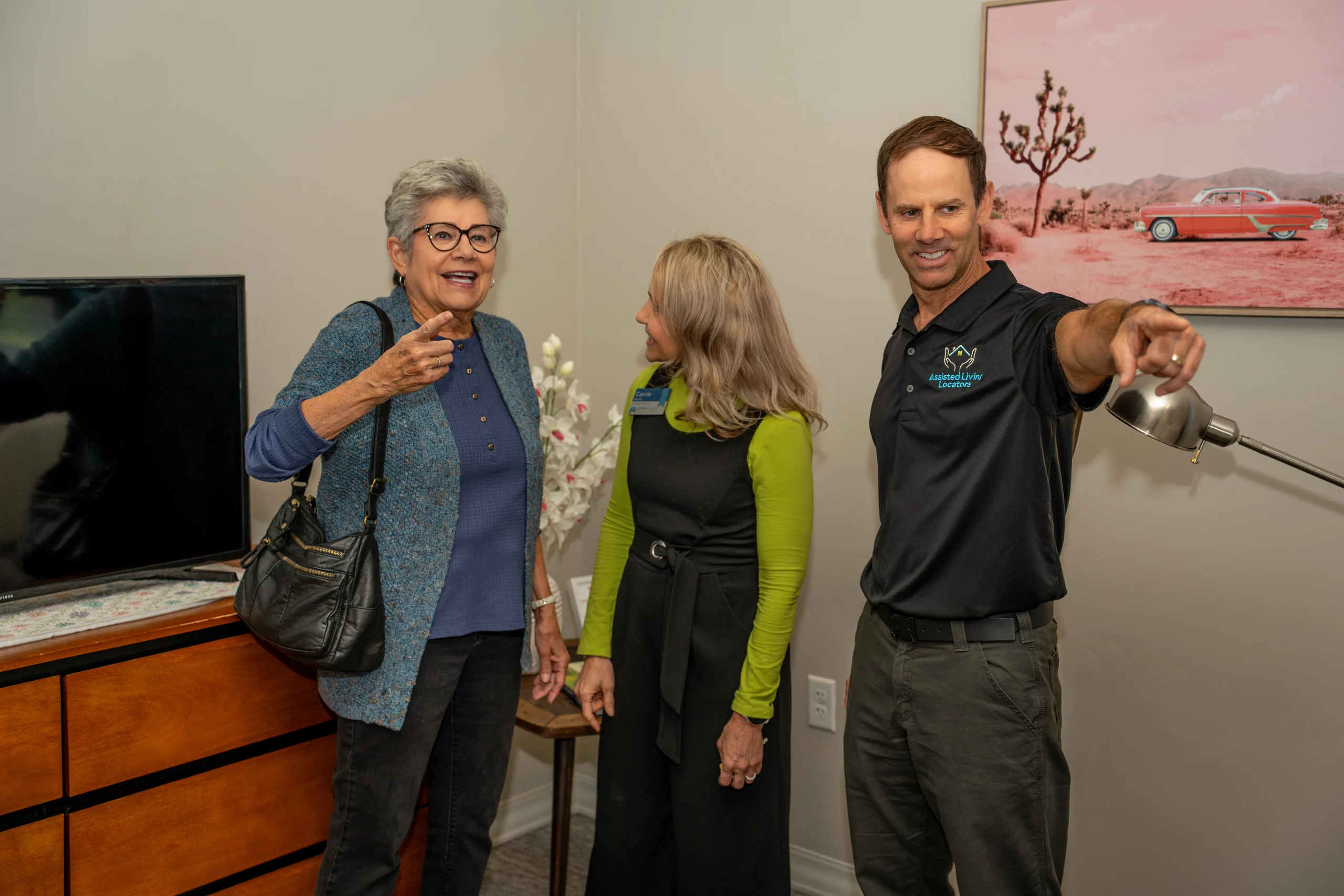 Three people standing in a living room, smiling and chatting—an older woman with glasses pointing ahead, a woman in a green top looking at her, and a man in a black polo pointing across the room, possibly giving a tour.